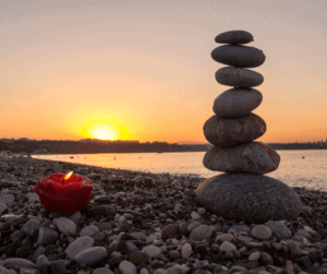 Candle with flame, pile of rocks balanced on each other, on the seashore with a sunset in the background