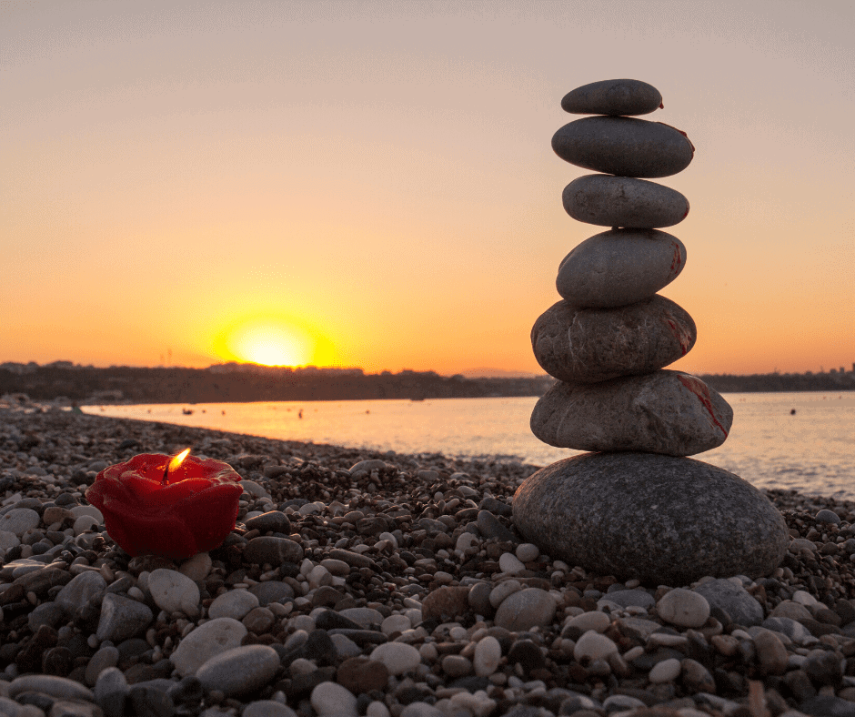 Candle with flame, pile of rocks balanced on each other, on the seashore with a sunset in the background to reflect holistic balance for body mind and spirit