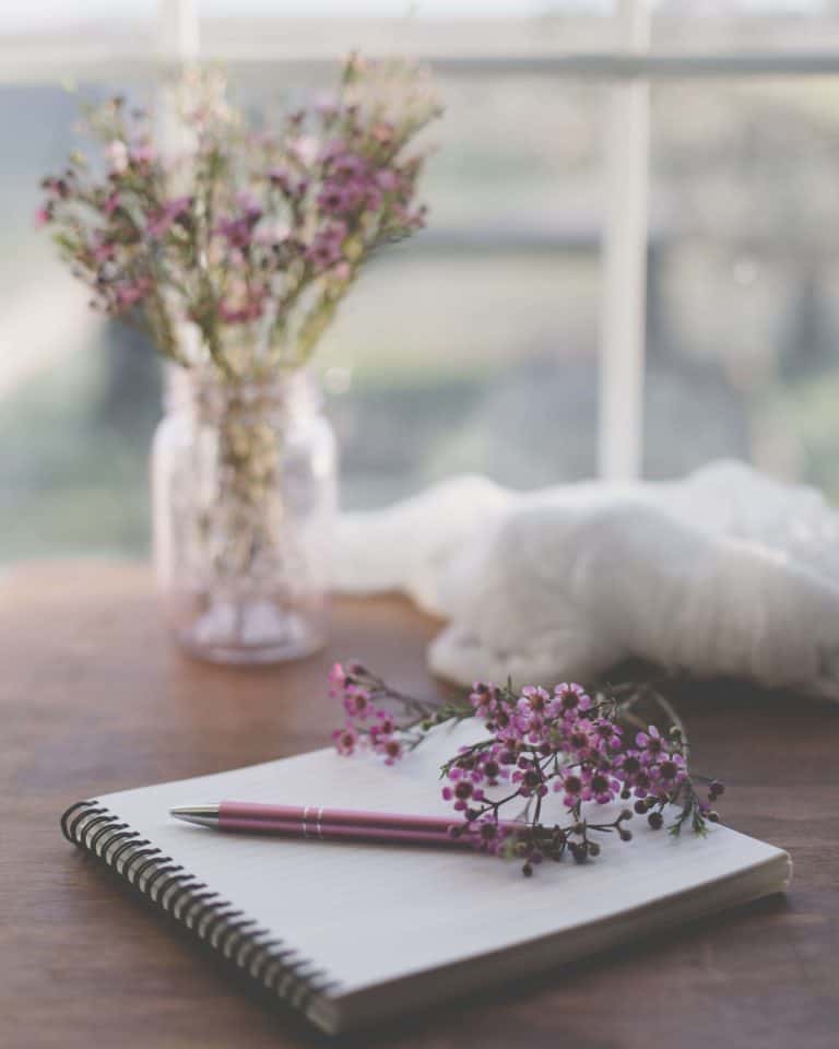 create a journaling habit requires only a pen and some paper. A notebook with a pen and vase of flowers on a desk top.