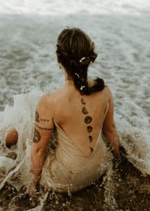 Woman with a moon phase tattoo down the center of her back, sitting in the tide on a beach.
