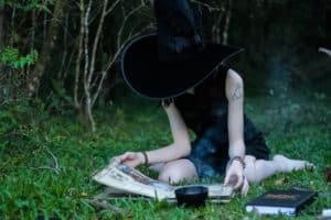 A sacred year journey for the solitary green witch might look like this woman dressed in black wearing a witch hat, seated on a blanket in the grass studying from a book.
