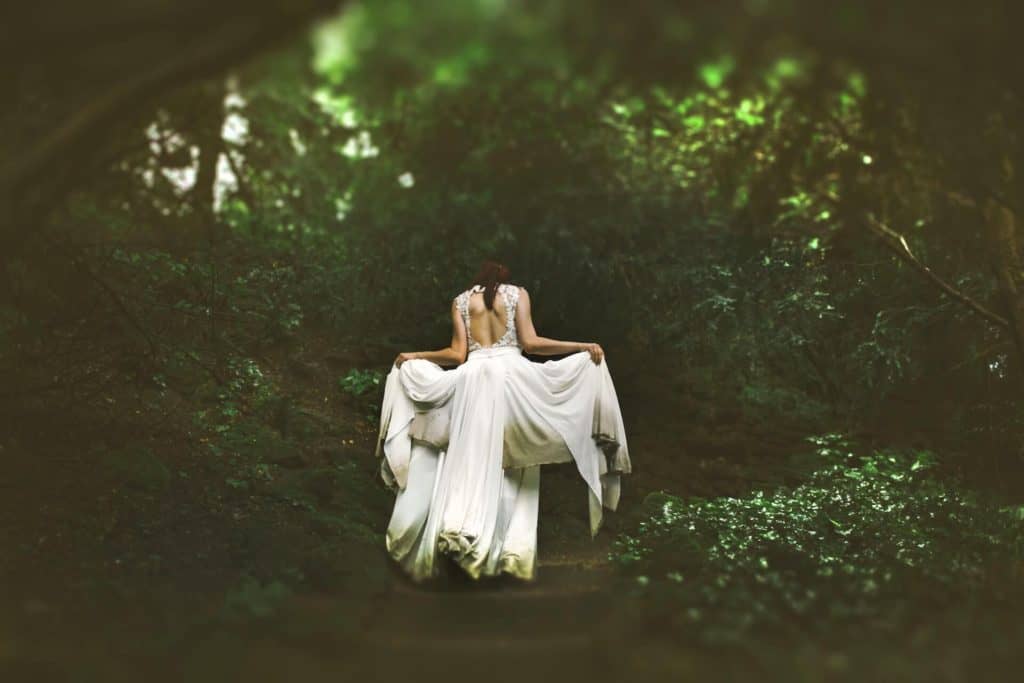 Green witch woman in a long white gown facing away from the viewer and going into the forest. Ravynwylde Waitlist
