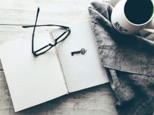 Image shows a journal laying open with a skeleton key and a pair of glasses beside it, ready for journaling. A coffee cup with a cow is to the side on a gray napkin.