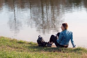 girl sitting beside a lake, with her back to us and a backpack at her feet. Listening with headphones. 