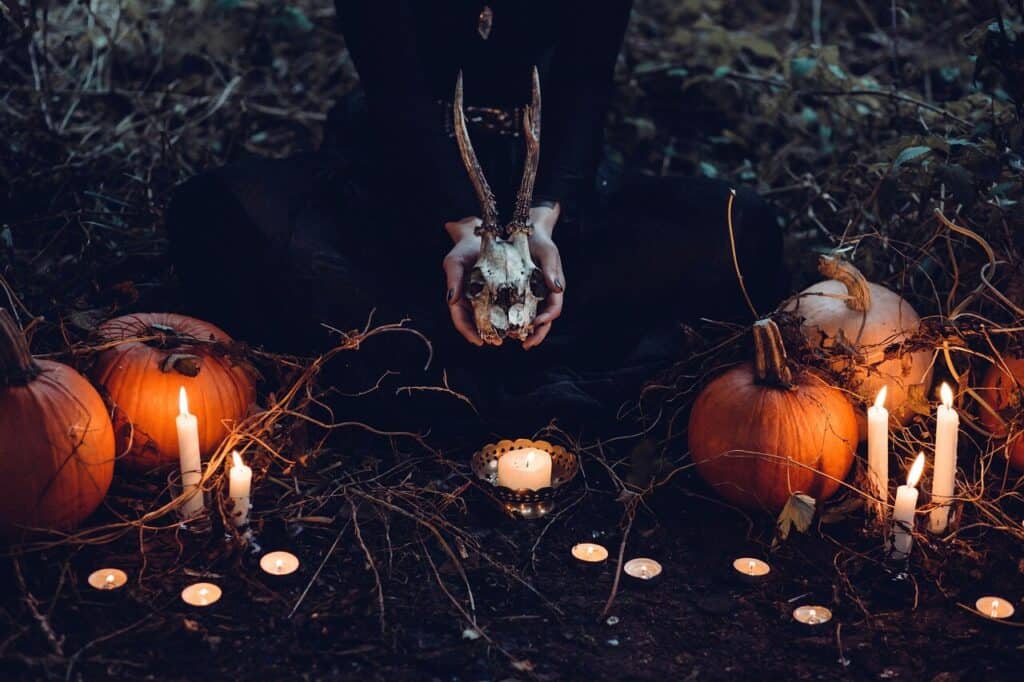 image depicts an altar for honoring the ancestors with a person in black, kneeling, face not visible holding a deer skull with antlers and surrounded by lit candles and orange pumpkins in a darkened image.