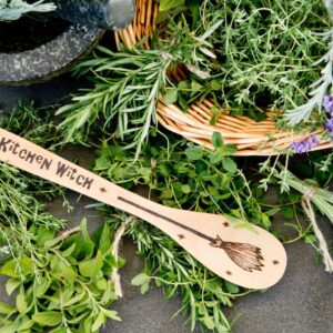 wooden spoon that says kitchen witch and has a broom engraved on it surrounded by fresh herbs