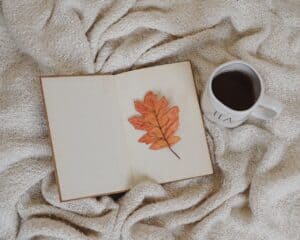open blank notebook with a single orange leaf sitting on the page with a cup of coffee nearby to represent creating a book of seasons for the green witch 