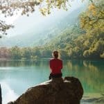 woman wearing red jacket with hair in an up do sitting on a boulder practicing meditation in a woodland setting near a lake