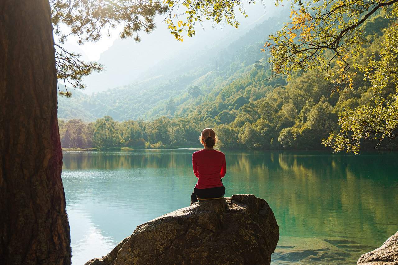 woman wearing red jacket with hair in an up do sitting on a boulder practicing meditation in a woodland setting near a lake