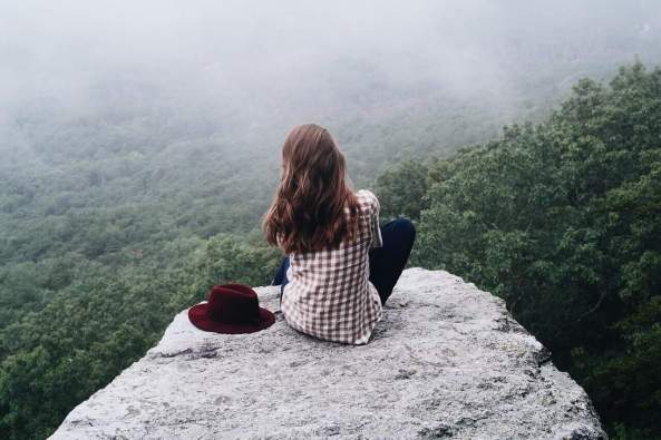 Woman with long dark hair seated on a rock outcropping overlooking a valley below. Nature connection through mindfulness. 