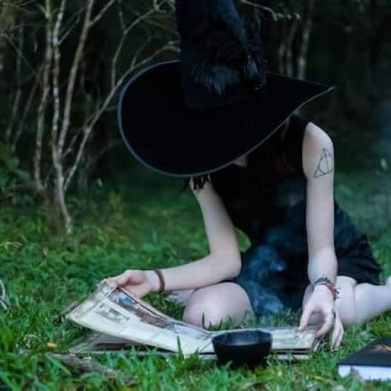 A sacred year journey for the solitary green witch might look like this woman dressed in black wearing a witch hat, seated on a blanket in the grass studying from a book.