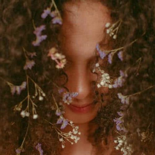 Ravynwylde - A Sacred Year Journey summer season image depicting a woman with long, dark curly hair and flowers woven into it, gazing toward her feet, which aren't visible.