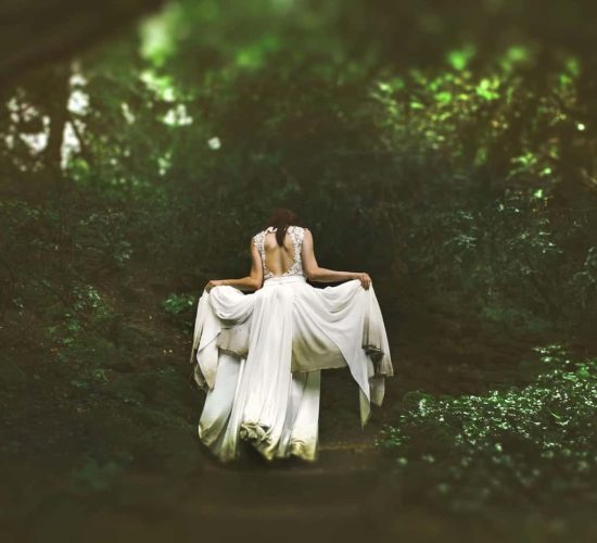 Green witch woman in a long white gown facing away from the viewer and going into the forest. Ravynwylde Waitlist