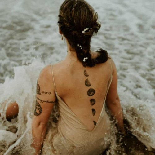 Woman with a moon phase tattoo down the center of her back, sitting in the tide on a beach.